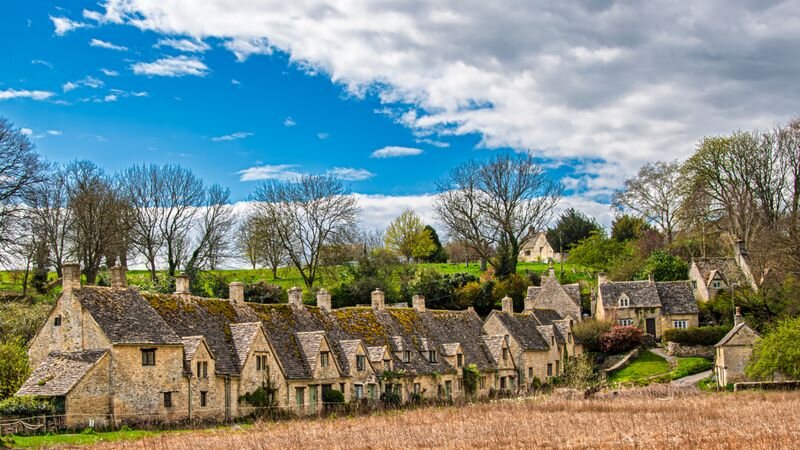 A row of golden stone cottages with steep gabled roofs along a village lane, framed by trees under a blue sky with wispy clouds