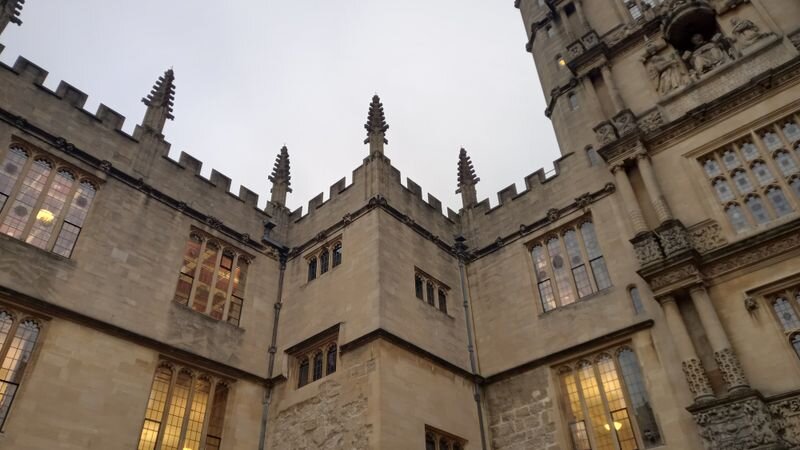 A Gothic university building facade with crenellated stone parapets, pinnacles, and lit mullioned windows at dusk