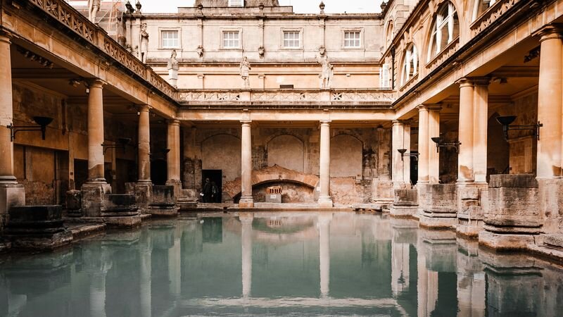 A Roman-style bath complex with stone colonnades and statues along the upper terrace, reflected in a still green pool