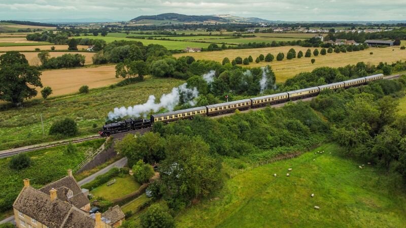 Aerial view of a vintage steam train pulling cream-and-black carriages through green countryside, past a stone farmhouse and grazing sheep